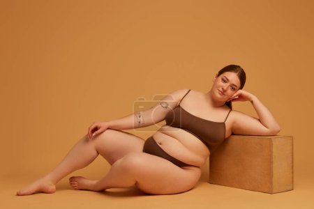 Photo for Equal beauty standards. Beautiful young girl with plus size body, wearing cotton underwear, posing against light brown studio background. Concept of natural beauty, body positivity, care, acceptance - Royalty Free Image
