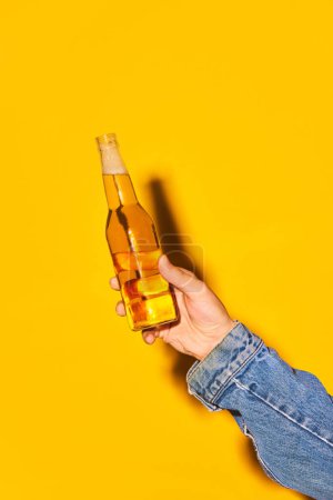 Téléchargez les photos : Main humaine tenant bouteille avec bière lager sur fond jaune vif. Concept de boisson alcoolisée et non alcoolisée, fête, vacances, bar, mix. Affiche. Espace de copie pour la publicité - en image libre de droit