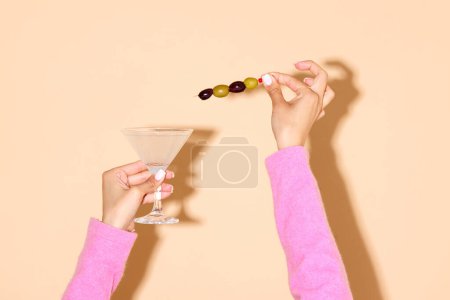 Téléchargez les photos : Main féminine tenant un verre de martini avec une olive verte sur un bâton sur fond clair. Concept de boisson alcoolisée et non alcoolisée, fête, vacances, bar, mix. Affiche. Espace de copie pour la publicité - en image libre de droit