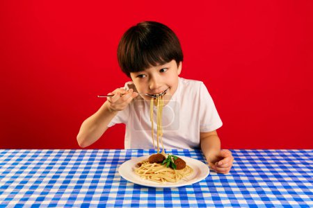 Photo for Little Korean boy, child sitting at table with checkered tablecloth and eating pasta with meatballs against red background. Concept of food, childhood, emotions, meal, menu, pop art - Royalty Free Image