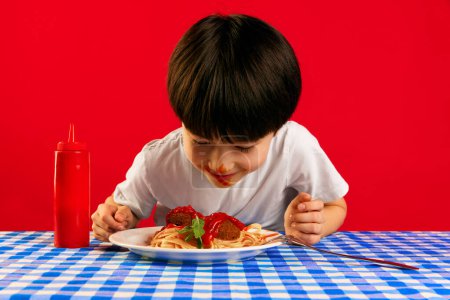 Foto de Niño, niño con la cara sucia de ketchup sentado en la mesa y comiendo deliciosos espaguetis con albóndigas y salsa de tomate sobre fondo rojo. Comida, infancia, emociones, comida, menú, concepto de arte pop - Imagen libre de derechos