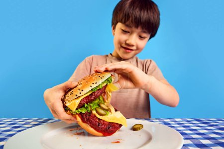 Foto de Pequeño coreano 7 orejas viejo niño comiendo hamburguesa jugosa gigante con verduras frescas y queso sobre fondo azul. Cena rápida. Concepto de comida, infancia, emociones, comida, menú, arte pop - Imagen libre de derechos