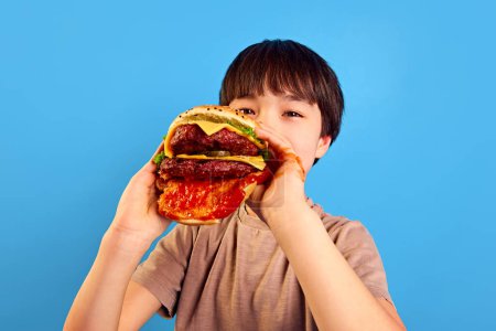 Foto de Niño, niño sosteniendo hamburguesa fresca gigante y jugosa con carne, salsa de tomate y queso, comiendo con una cara encantadora sobre fondo azul. Concepto de comida, infancia, emociones, comida, menú, arte pop - Imagen libre de derechos