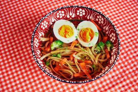 Téléchargez les photos : Soupe de nouilles ramen de style asiatique avec des légumes et des œufs parfaitement cuits, servie dans un bol décoratif sur une nappe à carreaux rouges. Concept de nourriture, repas, menu, pop art, cuisine japonaise - en image libre de droit
