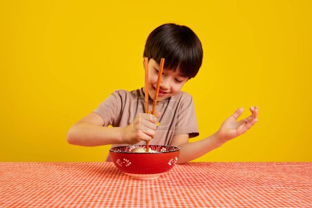 Téléchargez les photos : Garçon souriant assis à table et avec une assiette de soupe ramen et apprenant à manger avec des baguettes sur fond jaune. Concept de nourriture, enfance, émotions, repas japonais, menu, pop art - en image libre de droit