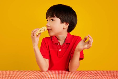 Photo for Korean boy in red polo shirt sitting at table and eating instant noodles against yellow background. Fast made dinner. Concept of food, childhood, emotions, meal, menu, pop art - Royalty Free Image