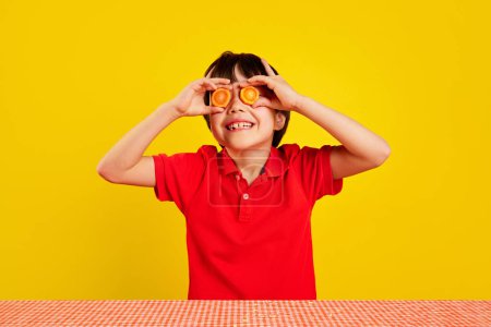 Foto de Niño alegre en polo rojo usando rodajas de zanahoria como gafas de mentira sobre fondo amarillo. Diversión creativa con la comida. Concepto de comida, infancia, emociones, comida, menú, arte pop - Imagen libre de derechos