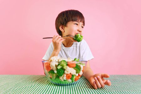 Photo for Little boy, child sitting at table and eating broccoli from a bowl full of fresh vegetables against pink background. Concept of food, healthy eating, childhood, emotions, meal, menu, pop art - Royalty Free Image