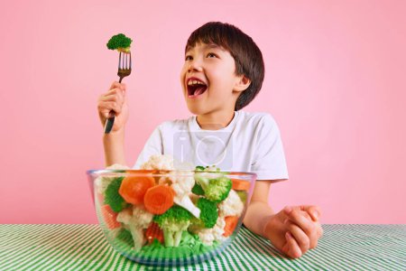 Photo for Playful and healthy. Smiling boy, kid raising for with broccoli, sitting at table with bowl full of boiled vegetables on pink background. Concept of food, healthy eating, childhood, emotions, pop art - Royalty Free Image