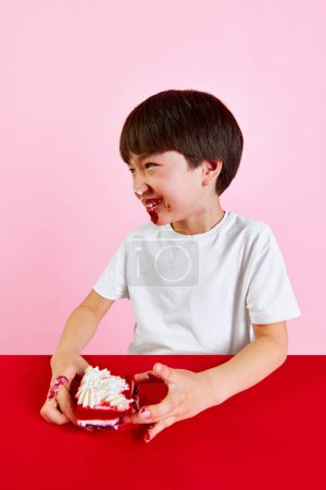 Téléchargez les photos : Petit garçon coréen souriant assis avec le visage sale dans la crème de gâteau, manger avec plaisir gâteau de velours rouge sur fond rose. Concept de nourriture, enfance, émotions, repas, menu, pop art - en image libre de droit