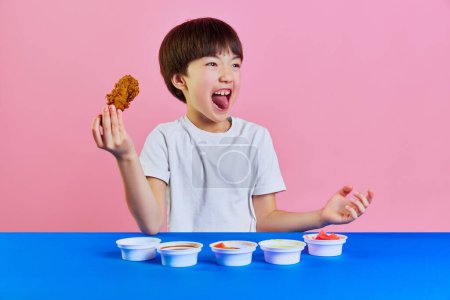 Photo for Emotional little Korean boy in white t-shirt eating spicy chicken, nuggets with different sauces against pink background. Concept of fast food, childhood, emotions, meal, menu, pop art - Royalty Free Image