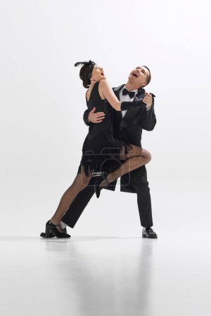 Foto de Joven pareja elegante en trajes negros vintage bailando alegremente Charleston con emociones expresivas aisladas sobre fondo blanco del estudio. Concepto de arte, retro y vintage, entretenimiento, 20s - Imagen libre de derechos