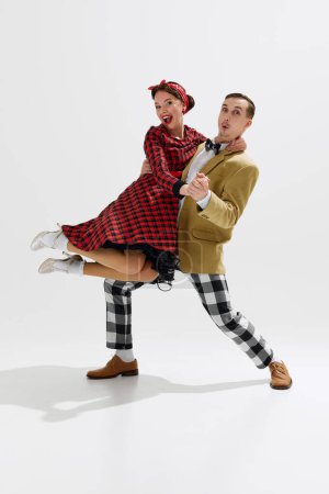 Téléchargez les photos : Amusant et énergique scène de danse rétro avec l'homme et la femme habillés dans la mode colorée des années 1950, partageant moment joyeux isolé surfond de studio blanc. Concept d'art, rétro et vintage, divertissement - en image libre de droit