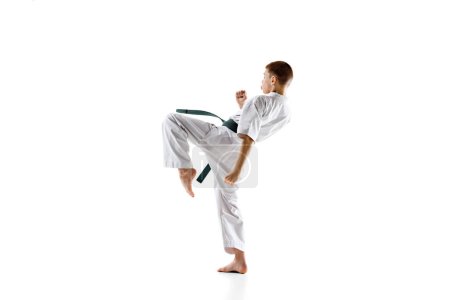 Foto de Estudiante de karate en kimono blanco y cinturón verde, en movimiento, realizando una poderosa patada durante la sesión de práctica, aislado sobre fondo blanco. Concepto de deporte, artes marciales, deporte de combate, estilo de vida activo - Imagen libre de derechos