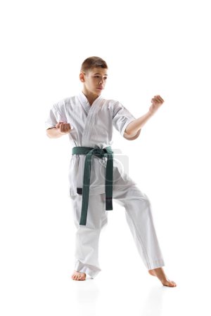 Teen boy in white karate and green belt demonstrating skills in stance, practicing isolated on white studio background. Concept of sport, martial arts, combat sport, healthy and active lifestyle