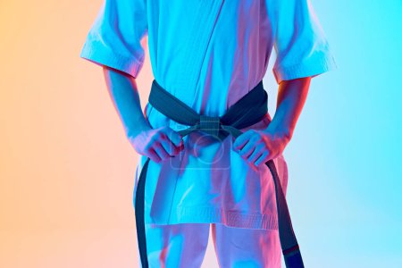 Foto de Imagen recortada de niño, atleta de karate en uniforme, kimono blanco y cinturón verde de pie contra el degradado fondo azul anaranjado en neón. Concepto de deporte, artes marciales, deporte de combate, estilo de vida activo - Imagen libre de derechos