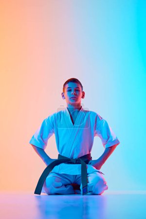 Foto de Niño adolescente, atleta de karate en uniforme, kimono blanco y cinturón verde sentado contra el degradado fondo azul anaranjado en neón. Concepto de deporte, artes marciales, deporte de combate, estilo de vida saludable y activo - Imagen libre de derechos