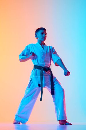 Foto de Karateka joven en una postura defensiva, adolescente practicando contra el degradado fondo azul anaranjado en luz de neón. Concepto de deporte, artes marciales, deporte de combate, estilo de vida saludable y activo - Imagen libre de derechos
