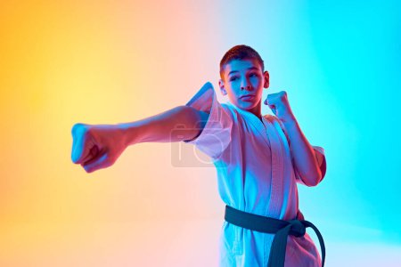Foto de Karateka enfocado, chico adolescente en uniforme practicando patadas, posturas contra el degradado fondo azul anaranjado en luz de neón. Concepto de deporte, artes marciales, deporte de combate, estilo de vida saludable y activo - Imagen libre de derechos