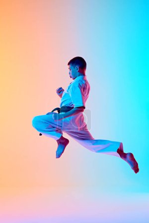 Young athlete engaged in karate training, jumping in dynamic pose against gradient orange blue background in neon light. Concept of sport, martial arts, combat sport, healthy and active lifestyle