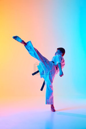 Foto de Imagen dinámica del adolescente, karateka en movimiento, mostrando una alta postura de patada contra el degradado fondo azul anaranjado en luz de neón. Concepto de deporte, artes marciales, deporte de combate, salud, estilo de vida activo - Imagen libre de derechos