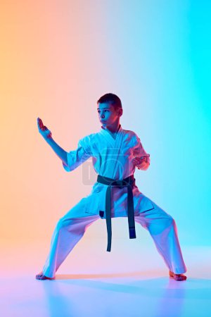 Young karate practitioner demonstrating kata stance in traditional white gi and green belt against gradient orange blue background in neon light. Concept of sport, martial arts, combat sport