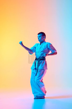 Foto de Postura de artes marciales dinámicas, adolescente en movimiento, entrenamiento contra el degradado fondo azul anaranjado en luz de neón. Concepto de deporte, artes marciales, deporte de combate, estilo de vida saludable y activo - Imagen libre de derechos
