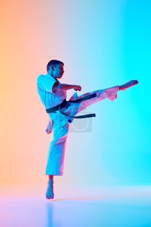 Full-length image of teen boy, karateka practicing high kick stance against gradient orange blue background in neon light. Concept of sport, martial arts, combat sport, healthy and active lifestyle