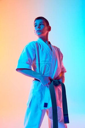 Foto de Niño adolescente, atleta de karate posando en kimono blanco con cinturón verde aislado sobre fondo blanco del estudio contra fondo azul anaranjado degradado en luz de neón. Concepto de deporte, artes marciales, deporte de combate - Imagen libre de derechos