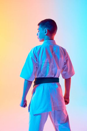 Photo for Back view image of teen boy, karate athlete posing in white kimono with green belt against gradient orange blue background in neon light. Concept of sport, martial arts, combat sport, active lifestyle - Royalty Free Image