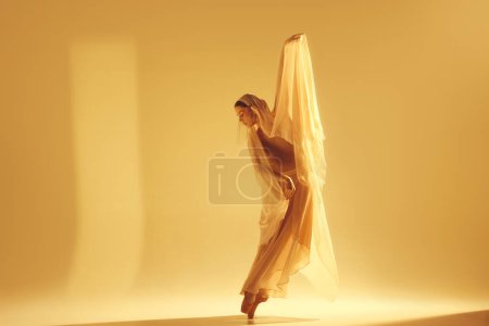 Foto de Danza de la luz. Bailarina de ballet femenina artística en vestido dorado que hace un rendimiento creativo elegante contra el fondo de color arena. Concepto de arte, danza clásica, belleza, moda, estética - Imagen libre de derechos