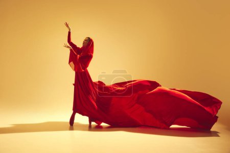 Foto de Pasión en movimiento. Hermosa mujer, bailarina en vestido rojo elegante que fluye haciendo un rendimiento poderoso contra el fondo de color arena. Concepto de arte, danza clásica, belleza y moda, estética - Imagen libre de derechos