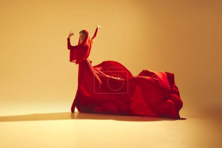 Téléchargez les photos : Élégance et passion. Danseuse artistique portant une robe fluide rouge et faisant des performances sur fond de couleur sable. Concept d'art, danse classique, beauté et mode, esthétique - en image libre de droit