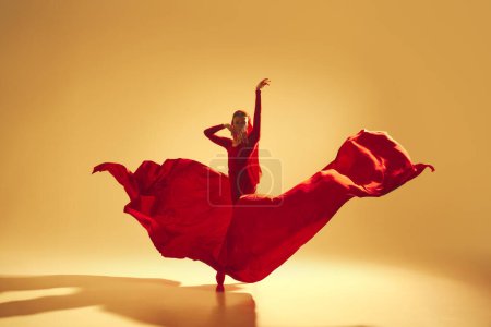 Foto de Actuación artística y profunda Mujer, bailarina femenina en vestido rojo fluido bailando sobre fondo de color dorado. Concepto de arte, danza clásica, belleza y moda, estética - Imagen libre de derechos