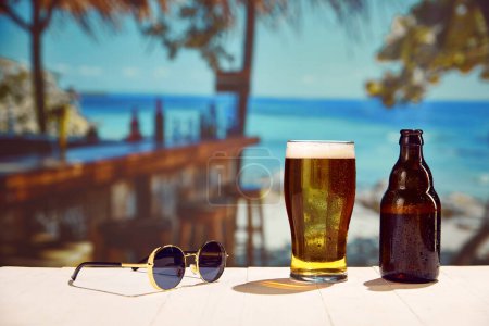 Foto de Escalofríos junto a la playa. Copa de cerveza helada, botella y gafas de sol mesa de madera, con fondo oceánico en el día cálido. Concepto de verano, vacaciones, relajación, viajes, tiempo libre - Imagen libre de derechos