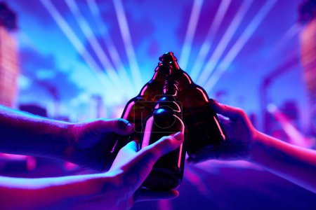 Photo for Cheers. Focus on beer bottles clinking against neon light background. People having fun at nightclub, attending party, concert. Concept of party, celebration, leisure activity, fun, night life - Royalty Free Image