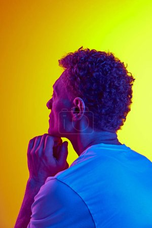 Photo for Man with curly hair, in casual t-shirt showing thoughtful expressing against yellow background with blue neon light. Concept of human emotions, facial expression, lifestyle, casual fashion - Royalty Free Image