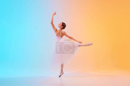 Téléchargez les photos : Photo dynamique de la danseuse de ballet étend son bras élégamment vêtue de tutu pâle dans la lumière au néon sur fond dégradé bleu-orange. Concept d'art, mouvement, fusion classique et moderne, beauté. Publicité - en image libre de droit