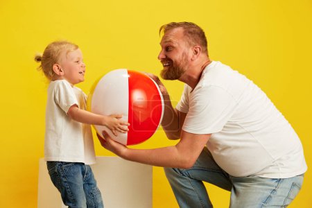 Photo for Man, father and his little boy share fun moment with beach ball, their expressions full of joy against yellow studio background. Concept of Happy Fathers day, family holidays celebration, parenthood. - Royalty Free Image