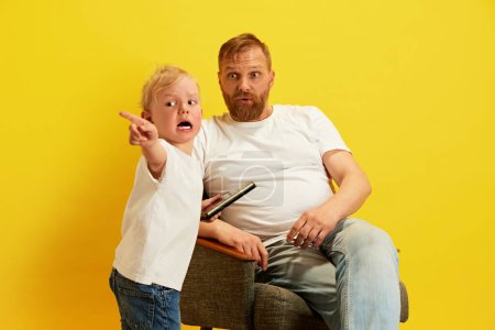 Photo for Father sits with surprised expression, holding remote control, while his son energetically points to distance against yellow studio background. Concept of Fathers day, family holidays, parenthood - Royalty Free Image