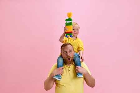 Photo for Cheerful father gives his son shoulder ride while he proudly holds up her colorful building block creation against pink studio background. Concept of Happy Fathers day, celebration, parenthood. Ad - Royalty Free Image