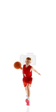 Téléchargez les photos : Vue de face du garçon motivé, joueur de basket en uniforme rouge en mouvement avec balle, jouant isolé sur fond de studio blanc. Concept de sport, enfance, école de sport, mode de vie actif - en image libre de droit