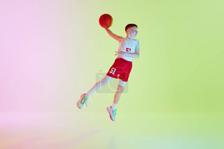 Téléchargez les photos : Image dynamique pleine longueur du bot en uniforme, joueur de basket-ball pratiquant, sautant avec la balle tenue dans une main contre fond de gradient dans la lumière du néon. Concept de sport, enfance, mode de vie actif - en image libre de droit