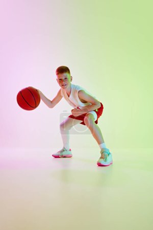 Téléchargez les photos : Un adolescent concentré, un joueur de basket en mouvement, une balle dribblante d'une main, un entraînement sur fond de gradient au néon. Concept de sport, enfance, école de sport, mode de vie actif - en image libre de droit