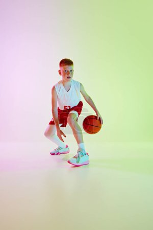 Téléchargez les photos : Il dégouline. Image complète du garçon en uniforme, jouant au basket-ball, s'entraînant contre un fond dégradé sous la lumière du néon. Concept de sport, enfance, école de sport, mode de vie actif - en image libre de droit
