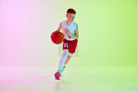 Téléchargez les photos : Joueur actif, adolescent garçon en uniforme jouant au basket-ball, en mouvement avec balle contre fond dégradé au néon. Concept de sport, enfance, école de sport, mode de vie actif - en image libre de droit