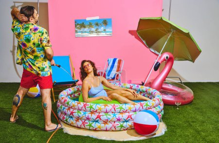 Foto de Rear view of man in tropical shirt and red shorts have fun spraying water at woman relaxing in inflatable pool. Concept of pop art, travelling, party, recreation, lifestyle, fashion and style. Ad - Imagen libre de derechos