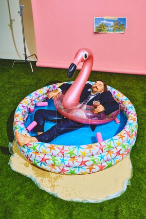 Foto de Young businessman in formal suit relaxing in kiddies swimming pool on flamingo inflatable-ring and drinks beer. Concept of pop art, party, recreation, lifestyle, fashion and style. - Imagen libre de derechos