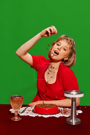 Téléchargez les photos : Quirky Meal Choice. Young woman in red dress eating from bowl of pet food against green background. Humorous dining. Concept of creative photography, pop art style, imagination, animal theme - en image libre de droit