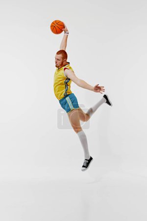 Slum Dunk. Ganzkörperbild eines jungen Mannes in gelbem Trikot und blauer Hose, Basketballspieler in Bewegung mit Ball auf weißem Hintergrund. Konzept aus Sport, Retro-Stil, aktivem Lebensstil, Hobby